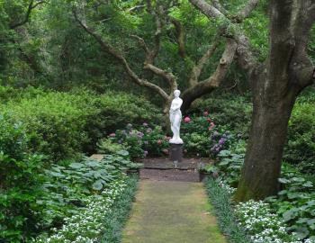 A white statue surrounded by trees, greenery, and a mix of pink and purple flowers