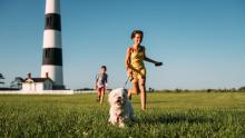 kids running thru grass with their dog; a lighthouse is in the background