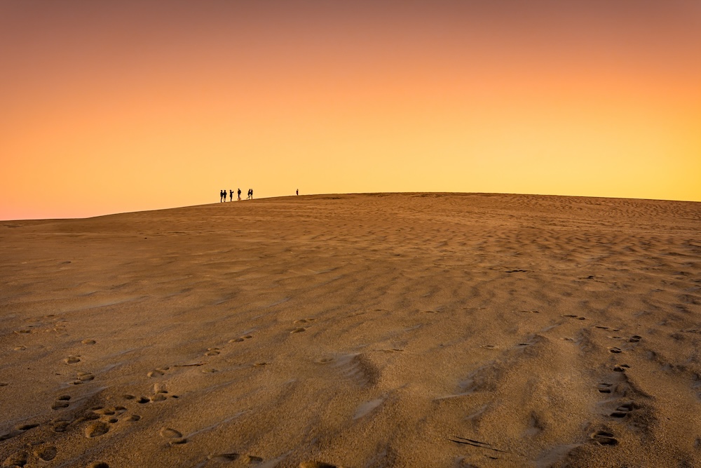 A group of people standing on top of a large sand dune at sunset