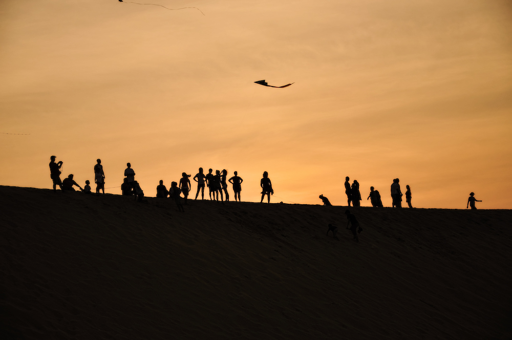 silhouetted people standing on top of Jockey's Ridge at sunset while flying kites